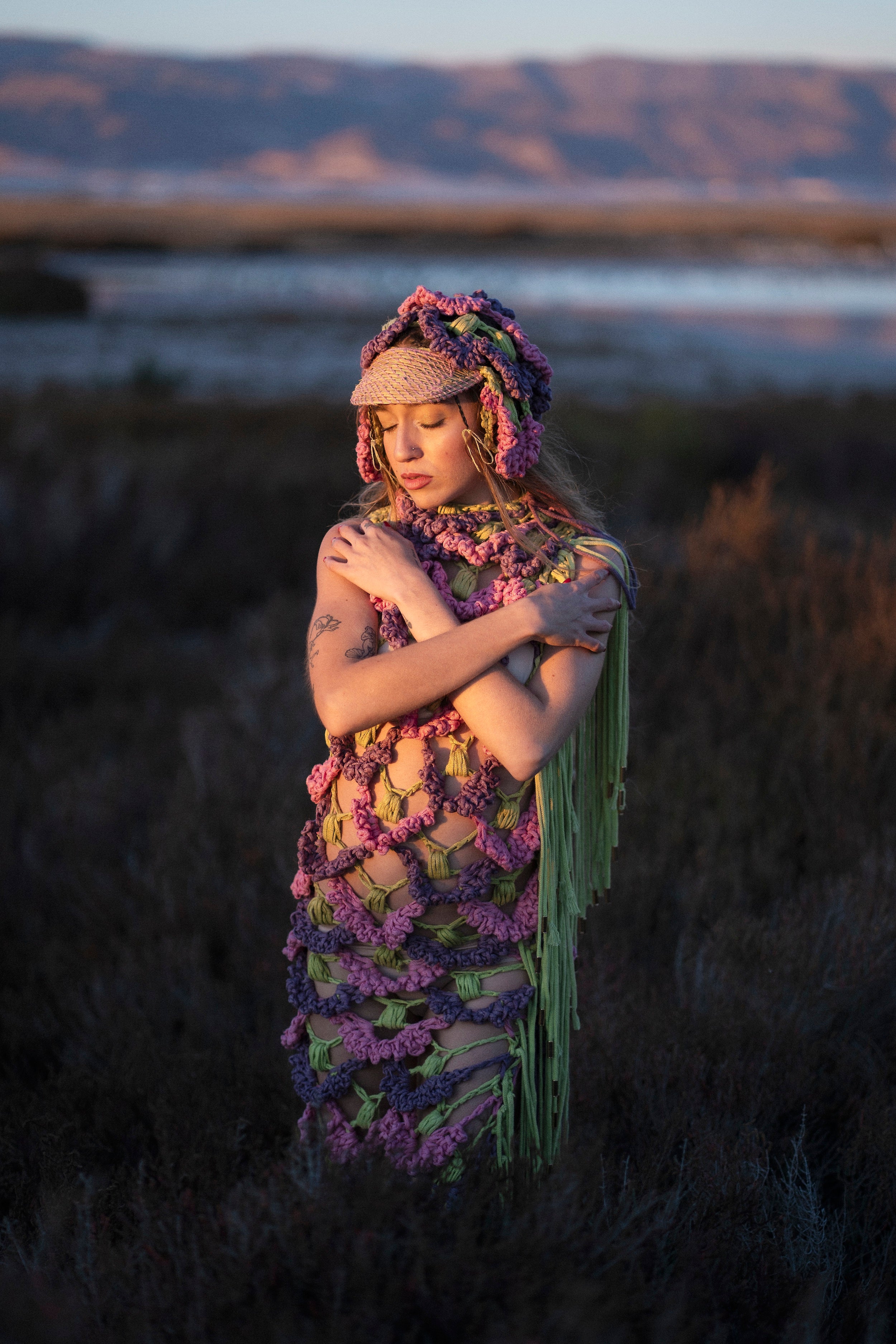 Front shot of the SYNTROPY dress. The pink, purple, and green dress features ACIEN's signature coronation crochet textile, made from pineapple and tender yarn, naturally dyed with plants, and adorned with green knitted cord fringing. Paired with a matching crochet cap.