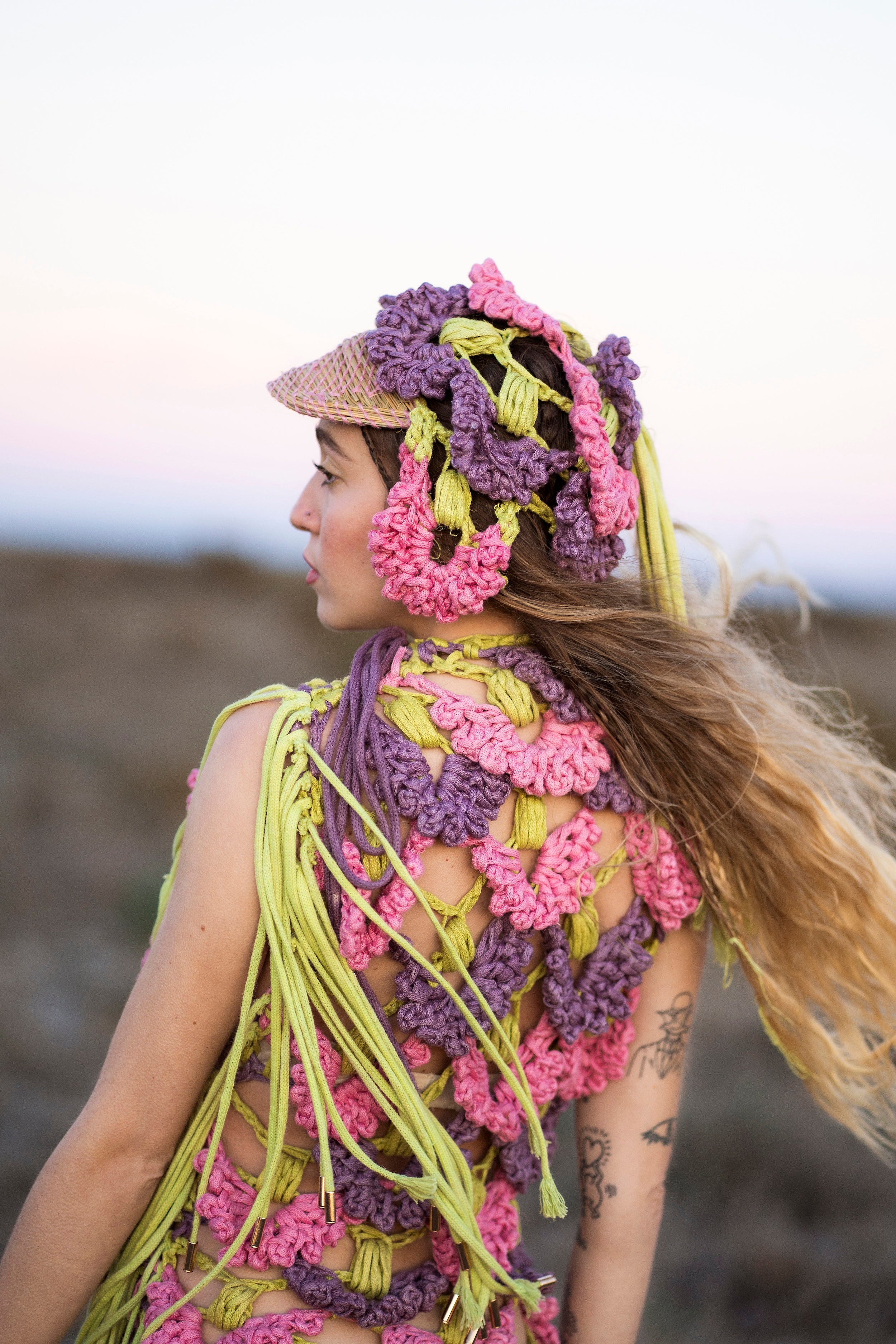 Close-up shot of the back of the SYNTROPY dress. The pink, purple, and green dress features ACIEN's signature coronation crochet textile, made from pineapple and tencel yarn, naturally dyed with plants, and adorned with green knitted cord fringing. Paired with a matching crochet cap.