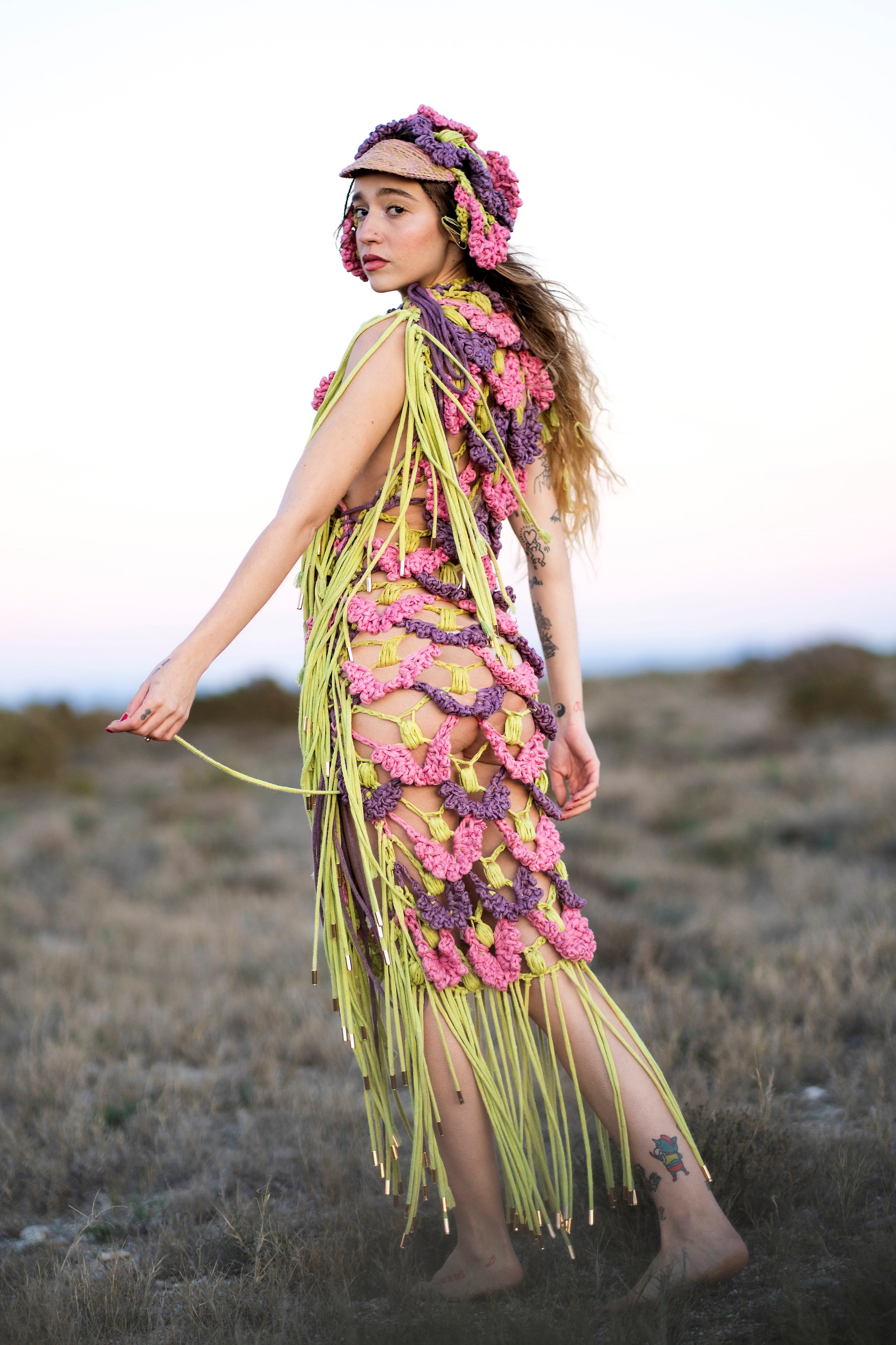 Wide shot of the back of SYNTROPY dress. The pink, purple, and green dress features ACIEN's signature tens crochet textile, made from pineapple and tencel yarn, naturally dyed with invasive plants, and adorned with green knitted cord fringing. Paired with a matching crochet cap.