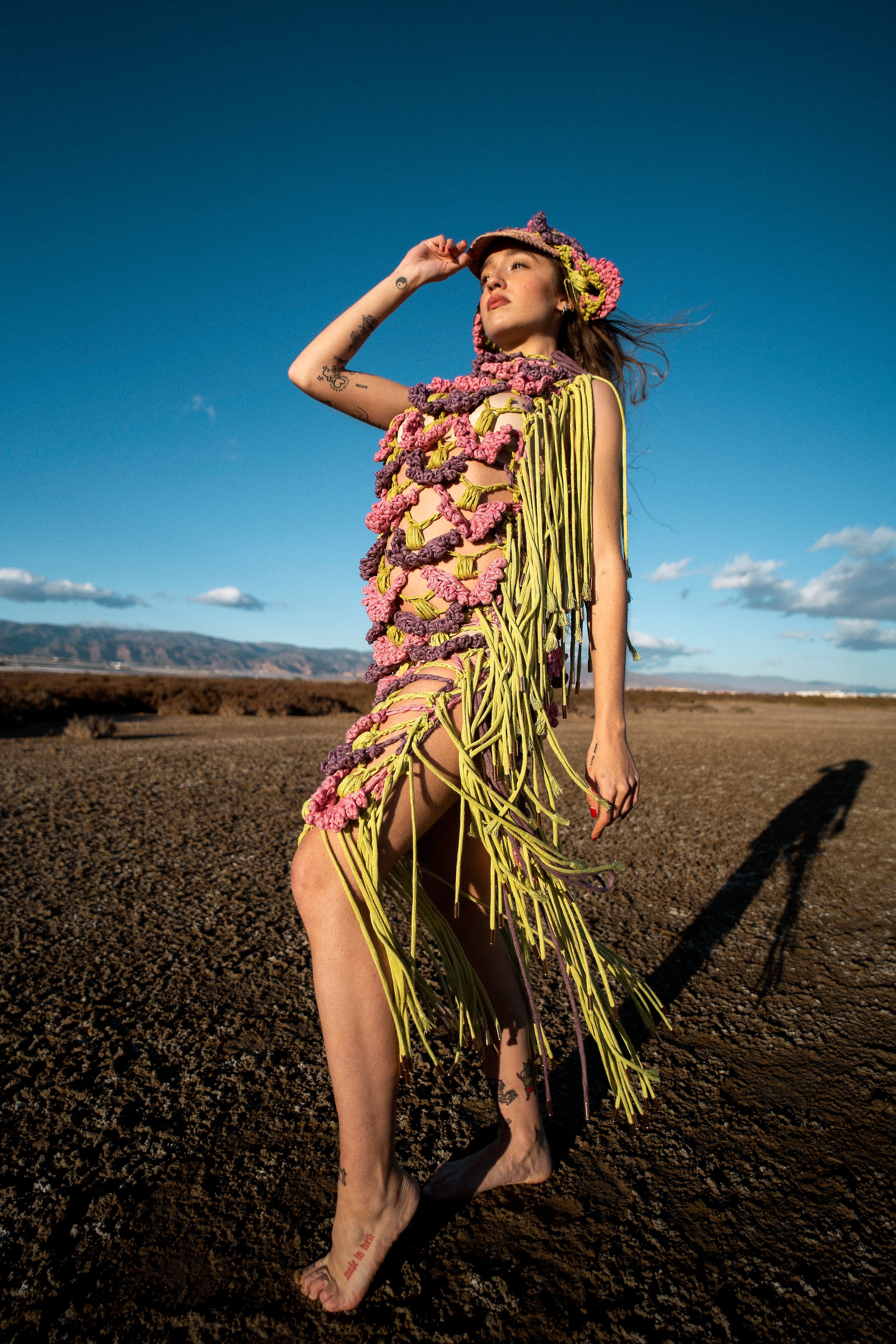 Side shot of the SYNTROPY dress. The pink, purple, and green dress features ACIEN's signature coronation crochet textile, made from pineapple and tencel yarn, naturally dyed with invasive plants, and adorned with green knitted cord fringing. Paired with a matching crochet cap.