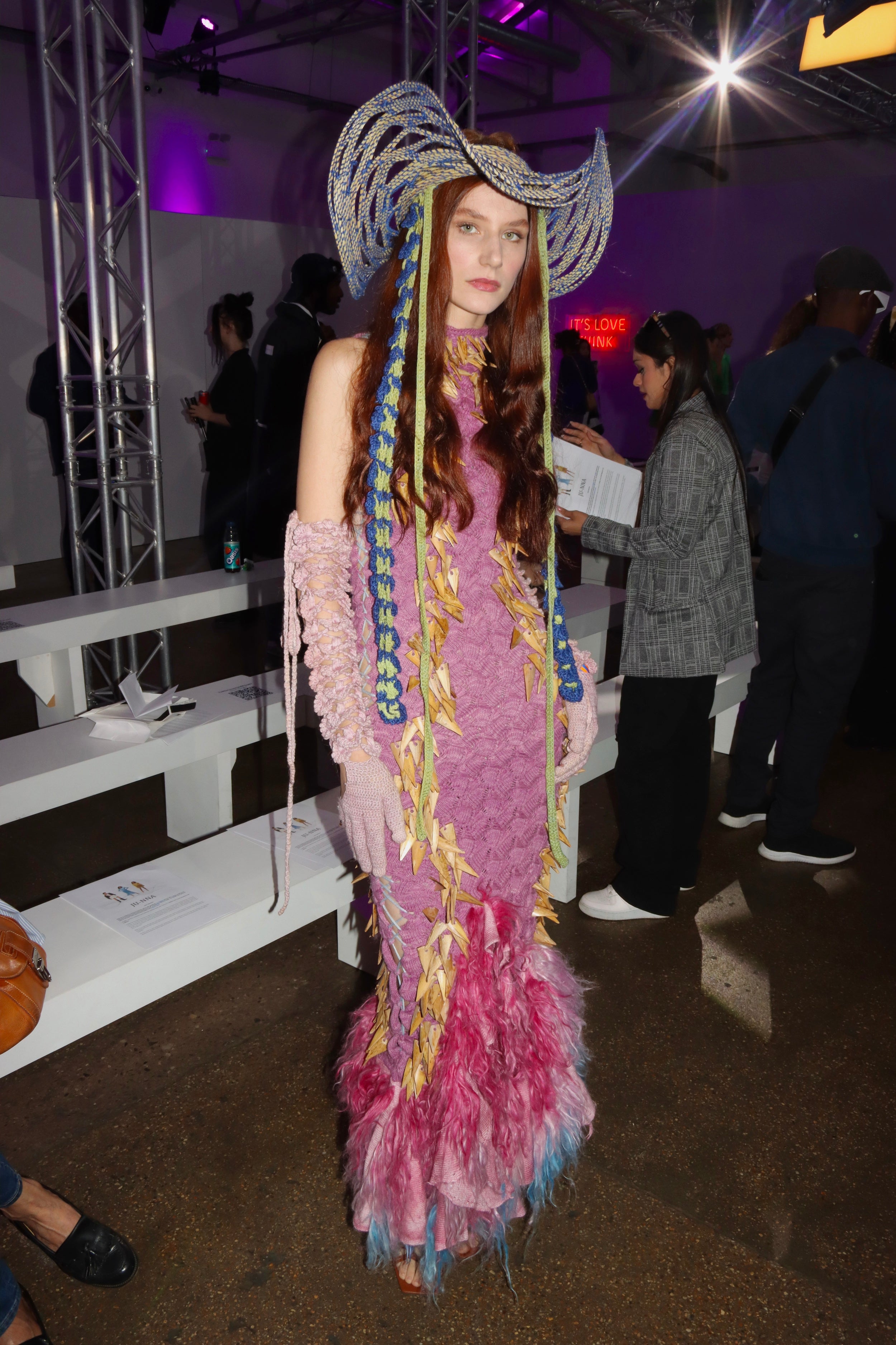 Tina Kolodiy wearing Look 3, TABERNAS, from REGENERATIVE FOLKLORE at London Fashion Week 2023. Pink knit dress made from nettle yarn, dyed from invasive plants adorned with hand cut Almería canes and pink fur detail at the bottom of the dress. Paired with  the TABERNAS hat, meticulously woven using ancestral techniques with dry branches sourced from the mountains of Southern Spain. Adorned with blue and green crocheted pineapple and tencel flower straps.